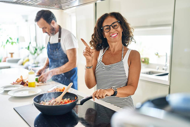 Pareja de mediana edad cocinando comida mediterránea en casa sonriendo feliz señalando con la mano y el dedo hacia un lado  - Foto, Imagen