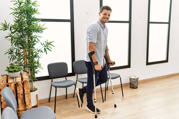 Young hispanic man smiling confident walking using crutches at clinic waiting room - 写真・画像