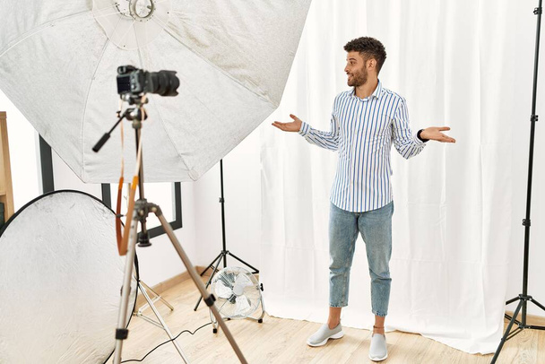 Arabischer junger Mann posiert lächelnd als Model im Fotostudio und zeigt mit beiden Händen geöffnete Handflächen, präsentiert und wirbt mit Vergleich und Balance  - Foto, Bild