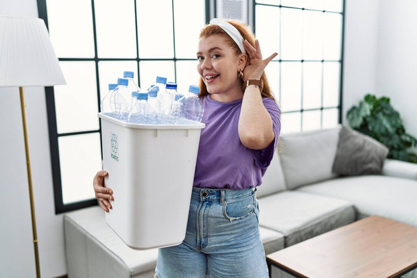 Young redhead woman holding recycling wastebasket with plastic bottles smiling with hand over ear listening an hearing to rumor or gossip. deafness concept.  - Photo, Image