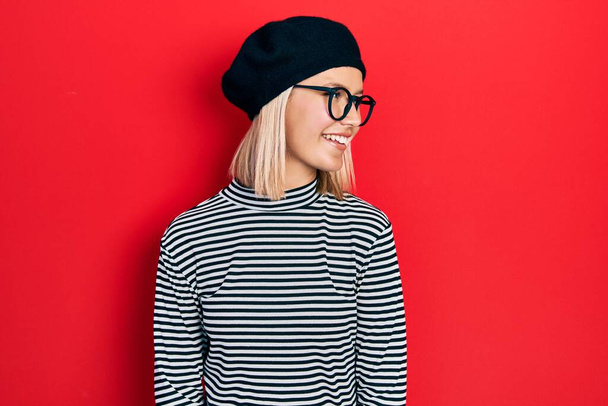 Beautiful blonde woman wearing french look with beret an glasses looking away to side with smile on face, natural expression. laughing confident.  - Photo, image