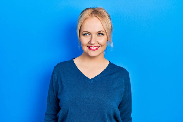 Jonge blonde vrouw draagt casual blauwe trui met een vrolijke en coole glimlach op haar gezicht. gelukkig persoon.  - Foto, afbeelding