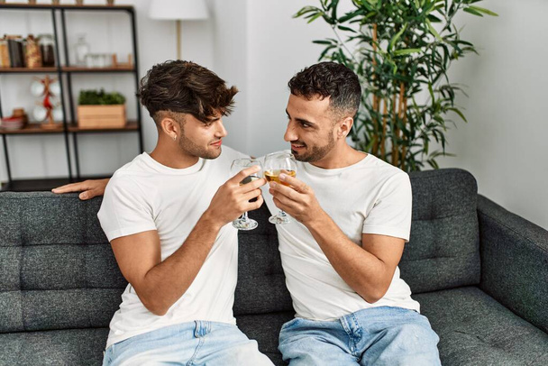 Dos hombres hispanos brindando en pareja con una copa de vino sentados en un sofá en casa - Foto, imagen