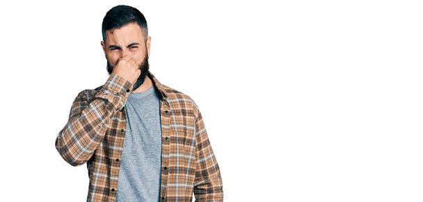 Homme hispanique avec barbe portant une chemise décontractée sentant quelque chose de puant et dégoûtant, odeur intolérable, retenant souffle avec les doigts sur le nez. mauvaise odeur  - Photo, image