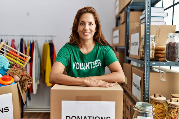 Young latin woman wearing volunteer uniform leaning on donations package at charity center - Photo, Image