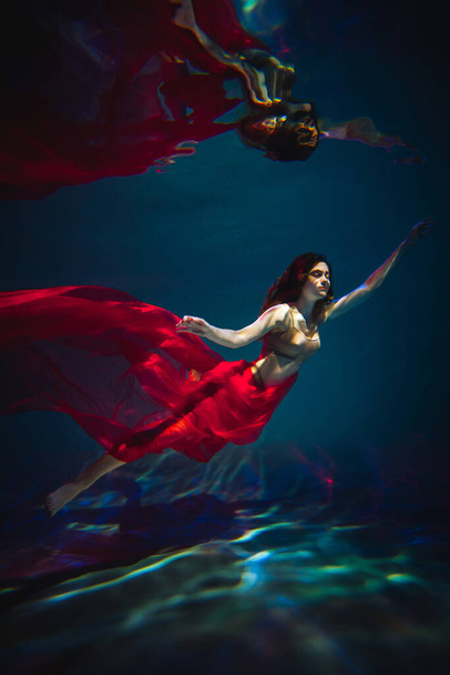 Έργο τέχνης. Υποβρύχια πλάνο ενός αθλητικού κοριτσιού με εσώρουχα με κόκκινο υλικό στην πισίνα με όμορφες ανταύγειες. - Φωτογραφία, εικόνα