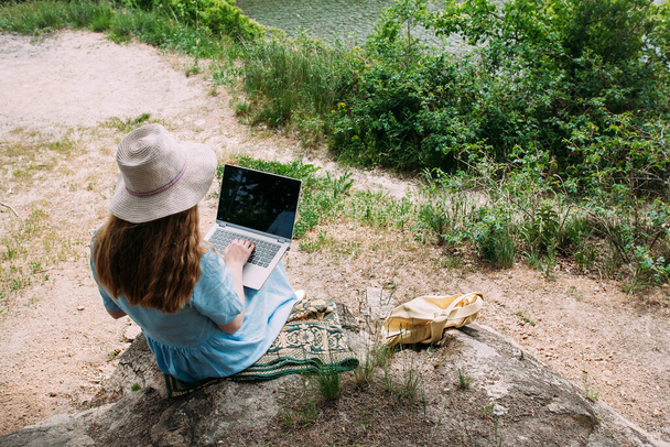 A young girl freelancer works with a laptop in nature. Dressed with a blue summer dress and a light hat. Sits on stones in a forested area. Nearby lies a beige leather bag. - Фото, зображення
