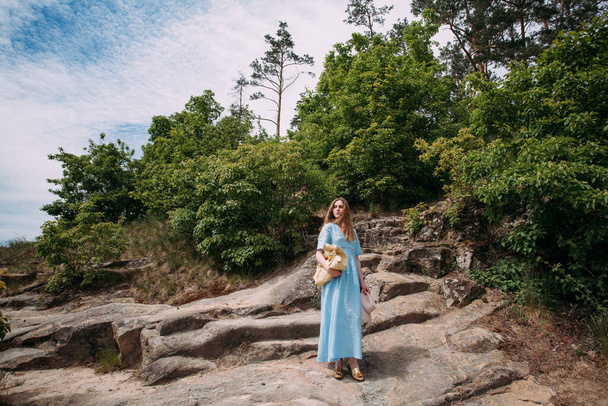 Een slank, jong meisje in een blauwe zonnejurk staat op stenen in de bergen, op een zonnige dag. In zijn handen draagt hij een roze hoed en een beige handtas. - Foto, afbeelding