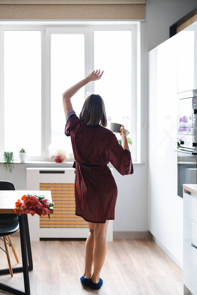 Happy Woman o kawę w domu, taniec i relaks, w kuchni w jedwabnym szlafroku, korzystających świeże kwiaty - Zdjęcie, obraz