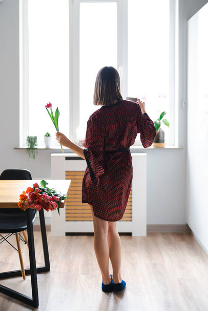 Mujer feliz tomando un café en casa, bailando y relajándose, en la cocina vistiendo una bata de seda, disfrutando de flores frescas - Foto, Imagen