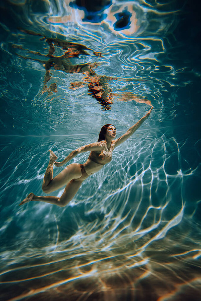 Έργο τέχνης. Υποβρύχια φωτογραφία ενός αθλητικού κοριτσιού με μπεζ εσώρουχα στην πισίνα με όμορφες ανταύγειες. - Φωτογραφία, εικόνα