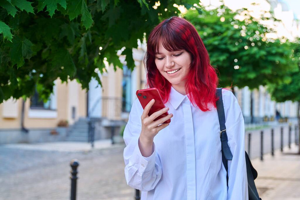 Beautiful smiling teenage girl looking at phone on city street. Female teenager with red dyed hair with backpack using smartphone. Lifestyle, technology, communication, youth, young people - Фото, зображення