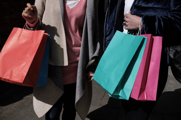 Κοντινό πλάνο δύο γυναικών που παίρνουν πολύχρωμες τσάντες για ψώνια. Διαφορετικά χρώματα, ηλιόλουστο πρωινό. Έννοια του καταναλωτισμού. - Φωτογραφία, εικόνα