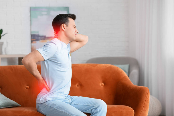 Молодой человек страдает от болей в спине, сидя дома на диване - Фото, изображение