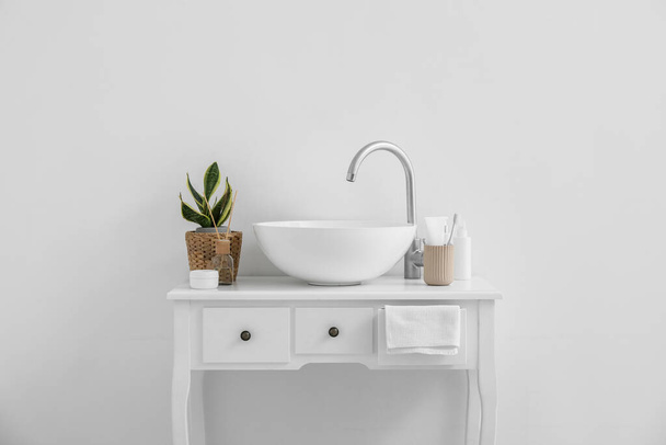 Table with sink, bath accessories and houseplant near light wall - Foto, afbeelding
