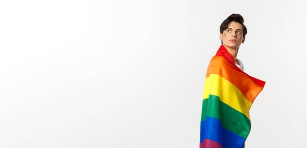 Bild eines nachdenklichen jungen schwulen Mannes mit lgbtq-Fahne, Kopf oben links drehen, Logo anstarren, vor weißem Hintergrund stehen. - Foto, Bild