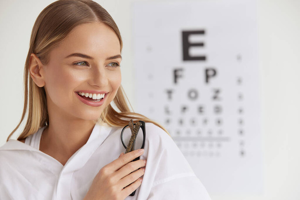 Optometry And Vision. Portrait Of Beautiful Smiling Girl Face At Ophthalmologist Office. Closeup Of Happy Young Woman With Healthy Eyes And Visual Eye Test Chart On Background. High Resolution Image - Фото, изображение