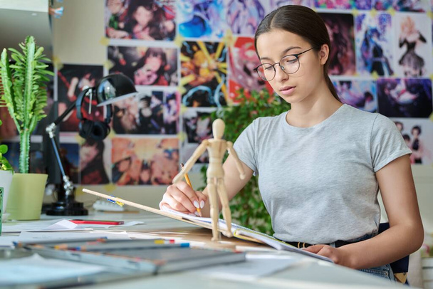Teenage creative girl artist drawing with a pencil, sitting at the table at home, using wooden mannequin. Creativity, school, hobby, leisure, lifestyle, adolescence, youth concept - Photo, image
