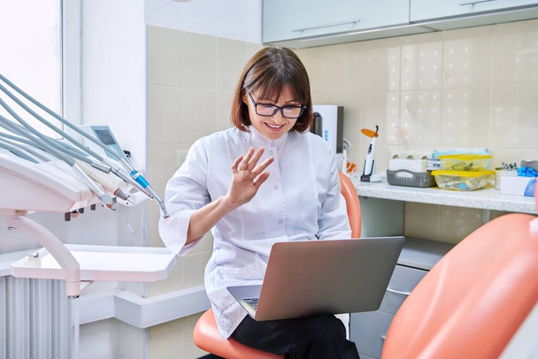 Konsultacja online, pomoc lekarza w klinice przy użyciu laptopa. Dentystka rozmawia machając ręką patrząc w laptopie w gabinecie dentystycznym. Wideokonferencja, wideokonferencja, serwis, koncepcja medycyny dentystycznej - Zdjęcie, obraz