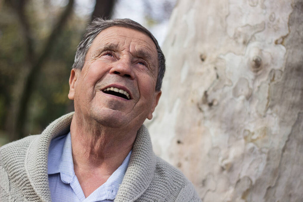 Πορτρέτο του ευτυχισμένου ηλικιωμένου άντρα χαμογελώντας, στο δημόσιο πάρκο, σε εξωτερικούς χώρους. Ο γέρος χαλαρώνει έξω και κοιτάει αλλού. Πορτρέτο του ηλικιωμένου άνδρα που απολαμβάνει σύνταξη - Φωτογραφία, εικόνα
