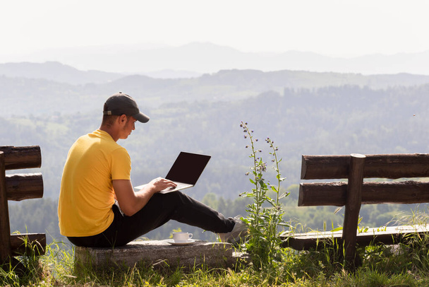 Jeune homme pigiste travaillant en ligne à l'aide d'un ordinateur portable, ordinateur portable de plein air dans la nature sur fond de paysage de montagne. Espace de copie pour la conception ou le texte sur l'écran noir d'ordinateur portable - Photo, image