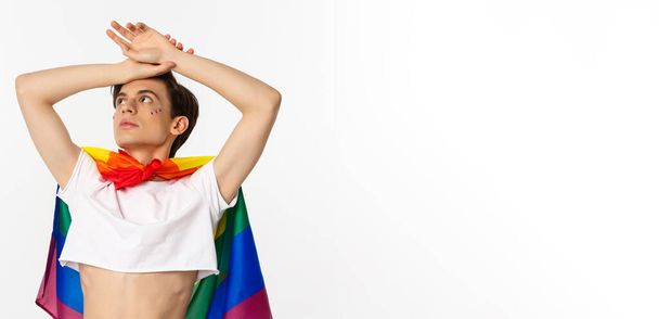 Schnappschuss eines sinnlichen androgynen Mannes mit bauchfreiem Oberteil und Regenbogenfahne, der mit erhobenen Händen in die linke obere Ecke blickt, weißer Hintergrund. - Foto, Bild