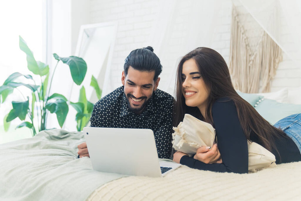 Relationship concept. Bright indoor shot of two people in a heterosexual relationship enjoying their free time by lying on bed and using social media on laptop. High quality photo - Photo, Image