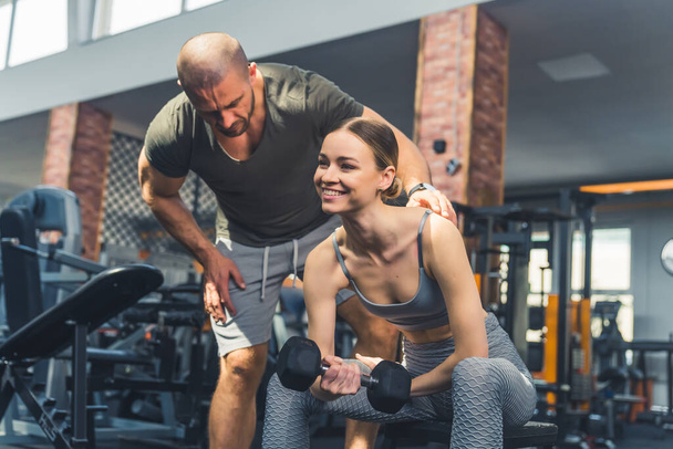 Gym concept. Caucasian woman lifting a dumbbell while sitting at a bench is smiling because now she sees she is a strong and powerful woman. Male personal trainer at the back. High quality photo - Foto, Bild