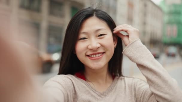 Söpö aasialainen tyttö ottaa omakuva osoittaa rauhan merkki kävellessään läpi kaupungin. Pitkäkarvainen positiivinen nainen tekee selfie ulkona - Materiaali, video
