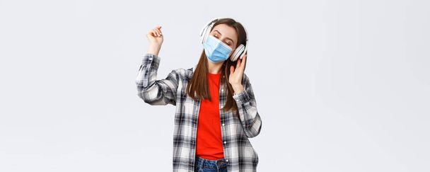 Social distancing, leisure and lifestyle on covid-19 outbreak, coronavirus concept. Carefree tender young woman carried away listening music in headphones, dancing with closed eyes in medical mask. - Photo, Image