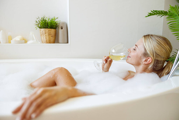 Hezká mladá běloška odpočívající ve vaně s bublinkami a popíjející bílé víno. Boční pohled. - Fotografie, Obrázek