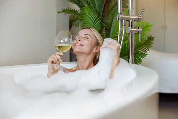 Attractive happy young woman enjoying bubbles foam and white wine in hot bathtub in spa. Relaxing. - Photo, Image