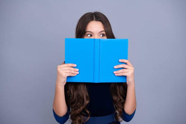 Schoolchild, teenage student girl hold book near eyes on gray isolated studio background. School and education concept. Back to school. Face hidden with book - Photo, image