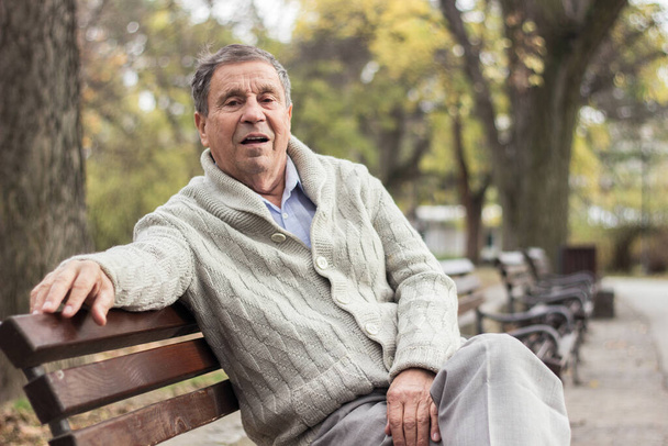Portré egy töprengő idős emberről, aki a padon ül, a nyilvános parkban, a szabadban. Az öregember a szabadban pihen és a kamerába néz. Idős nyugdíjas férfi portréja - Fotó, kép