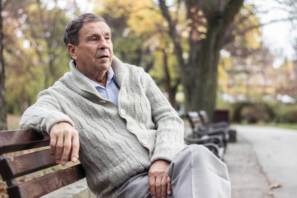 Portrét zamyšleného staršího muže sedícího venku na lavičce ve veřejném parku. Stařec odpočívá venku a dívá se jinam. Portrét staršího muže v důchodu - Fotografie, Obrázek