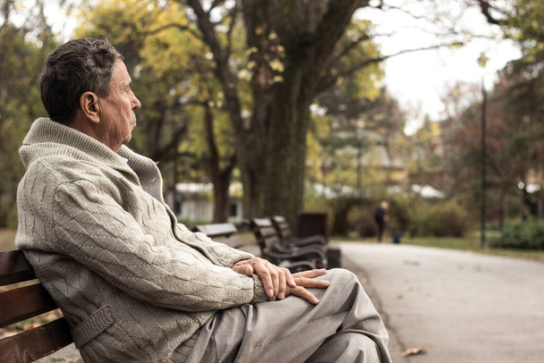 Портрет задумчивого пожилого человека, сидящего на скамейке, в общественном парке, на улице. Старик отдыхает на свежем воздухе и смотрит в сторону. Портрет пожилого человека, наслаждающегося пенсией - Фото, изображение