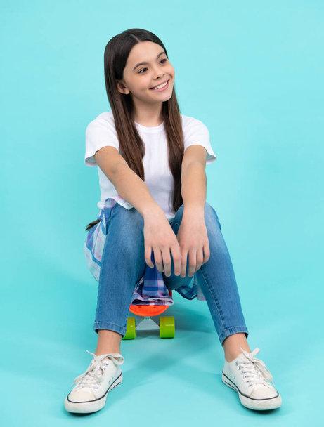 Happy vogue teenager. Fashionable confident teenage girl wearing trendy t-shirt and jeans sit on skateboard, posing on blue background. Fashion teenager, casual look - Foto, afbeelding