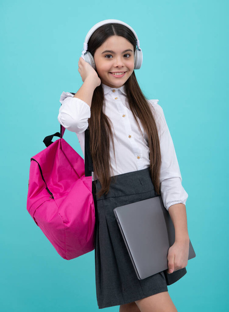 Okula dönelim. Okul üniformalı, çantalı, kulaklıklı ve dizüstü bilgisayarı olan bir kız öğrenci. İzole edilmiş mavi arka planda okul çocukları - Fotoğraf, Görsel