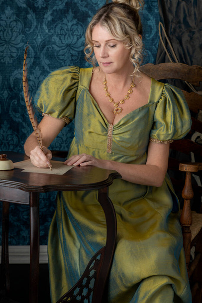 Una mujer rubia Regency vistiendo un vestido de seda verde y sentada en una pequeña mesa escribiendo una carta con una pluma - Foto, imagen
