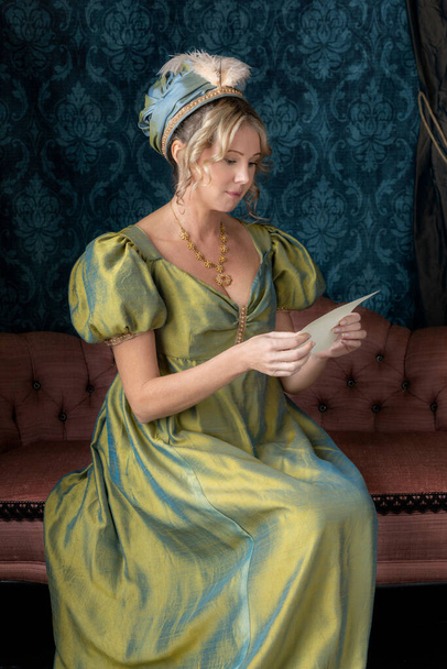 Eine blonde Regency-Frau in einem grünen Seidenkleid sitzt auf einem Sofa und hält einen Brief in der Hand - Foto, Bild