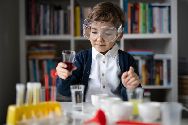 Jeden mały biały chłopiec naukowiec pięcioletni noszący okulary ochronne siedzący przy stole bawiący się sprzętem chemicznym zabawka wstępnie formująca eksperyment nauka i edukacja koncepcja widok z przodu - Zdjęcie, obraz