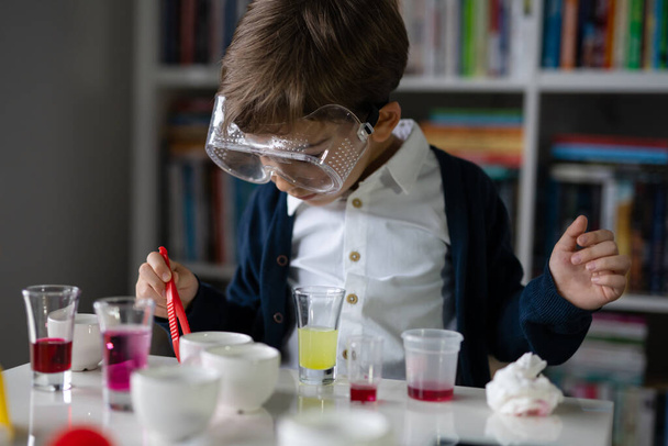 Jeden mały biały chłopiec naukowiec pięcioletni noszący okulary ochronne siedzący przy stole bawiący się sprzętem chemicznym zabawka wstępnie formująca eksperyment nauka i edukacja koncepcja widok z przodu - Zdjęcie, obraz