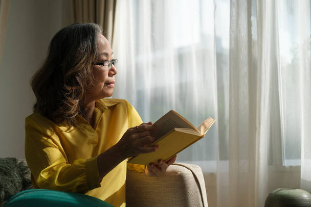 Gelukkige oudere vrouw neemt een pauze van het lezen terwijl ze door het raam kijkt. Pensioenconcept. - Foto, afbeelding