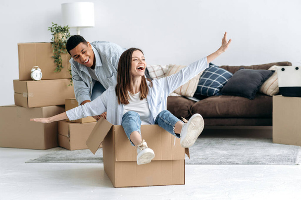 New home, relocation. Happy mixed race young family couple, bought their own house, they having fun on moving day, joyful wife sitting in cardboard box and husband push her, they laughing together - Photo, Image