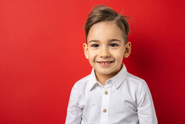 One boy male child five years old in front of red background wall wearing white shirt looking to the camera confident copy space waist up happy smile - Foto, imagen