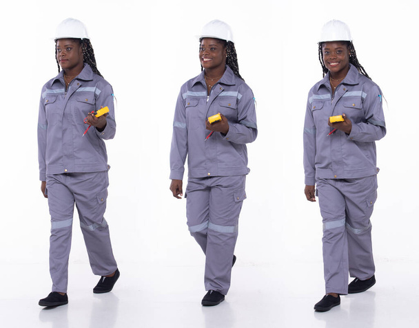 Młoda Afroamerykanka, elektryk przemysłowy, idąca do przodu w prawo, ubrana w formalne spodnie marynarskie i buty. Inżynier kobieta nosić tabletki internetowe na białym tle izolowane - Zdjęcie, obraz