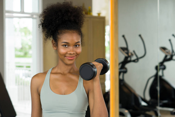 Jonge krullende zwarte haarvrouw genieten van vakantie in fitnesscentrum. Tillen lange halter met een hand tijdens het glimlachen. - Foto, afbeelding