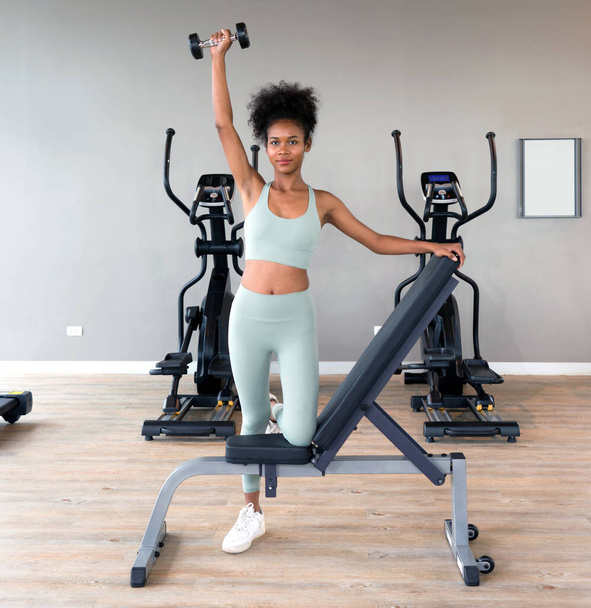 Νέοι σγουρά μαύρα μαλλιά γυναίκα απολαμβάνουν τις διακοπές στο γυμναστήριο. Σηκώνει το κουδούνι με το ένα χέρι, ενώ ξεκουράζει το γόνατό της στον πάγκο πολλαπλών λειτουργιών. Υπάρχουν μηχανήματα καρδιο στο γυμναστήριο - Φωτογραφία, εικόνα