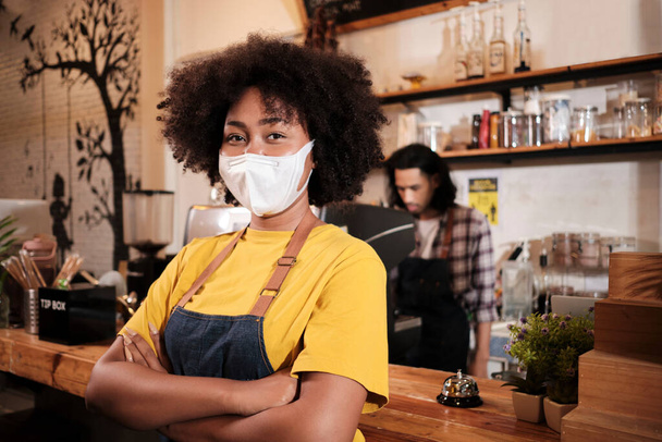 Retrato de una joven barista afroamericana con máscara facial mirando a cámara, brazos cruzados y trabaja con una sonrisa en un nuevo servicio de estilo de vida normal, negocio de pymes en cuarentena pandémica COVID19. - Foto, Imagen