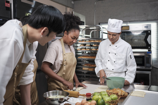 Cuisine course, senior male chef in cook uniform teaches young cooking class students to knead and roll pastry dough, prepare ingredients for bakery foods, fruit pies in stainless steel kitchen. - Fotó, kép